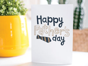 A greeting card is on a table top with a yellow plant pot and a green plant inside. The card features the words “Happy Father’s  Day” with a striped tie on the bottom of the words. 