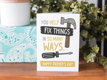 Load image into Gallery viewer, A greeting card is on a table top with a present in blue wrapping paper in the background. On top of the present is a candle and some greenery from a plant too. The card features the words  “You Help Fix Things in so Many Ways, Happy Father&#39;s Day” with an illustrated hammer and screwdriver around the words. 