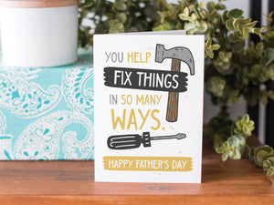 A greeting card is on a table top with a present in blue wrapping paper in the background. On top of the present is a candle and some greenery from a plant too. The card features the words  “You Help Fix Things in so Many Ways, Happy Father's Day” with an illustrated hammer and screwdriver around the words. 