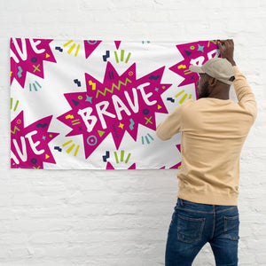 A man hanging a large flag on a white brick wall. The white flag features a pattern with the word "brave" featured inside a pink star-like shape with sketchy illustrations around the word. 