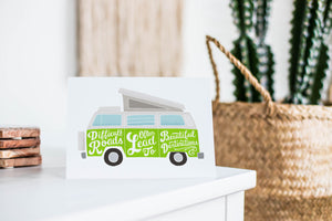 A greeting card featured standing up on a white tabletop with a framed photo of a succulent in the background and a stack of wooden coasters. There’s a woven basket in the background with a cactus inside. The card features the words “Difficult roads often lead to beautiful destinations” with an illustrated campervan.