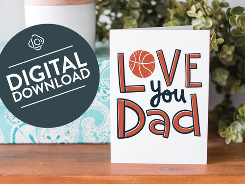 A greeting card is on a table top with a present in blue wrapping paper in the background. On top of the present is a candle and some greenery from a plant too. The card features the words  “Love you Dad” with an illustrated basketball as the “O” of love. The words 