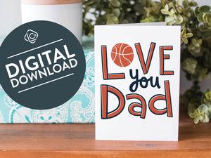 A greeting card is on a table top with a present in blue wrapping paper in the background. On top of the present is a candle and some greenery from a plant too. The card features the words  “Love you Dad” with an illustrated basketball as the “O” of love. The words "digital download" are featured in a circle on top of the image. 