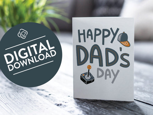 A greeting card featured on a black, wood coffee table. There’s a white planter in the background with a green plant. There’s also a gray sofa in the background with a white pillow. The card features the words “Happy Dad’s Day” with an illustrated game controller and hat. The words 