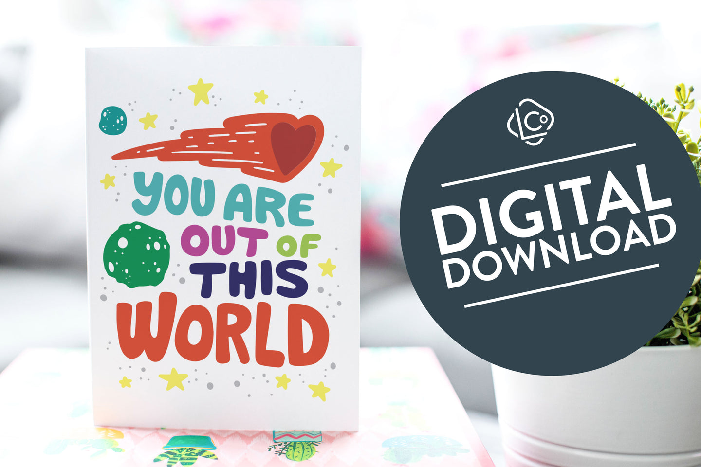 A greeting card is on a table top with a gift in pink wrapping paper. Next to the gift is a white plant pot with a green plant. The card features the words “You are out of this world” with space themed illustrations.. The words 