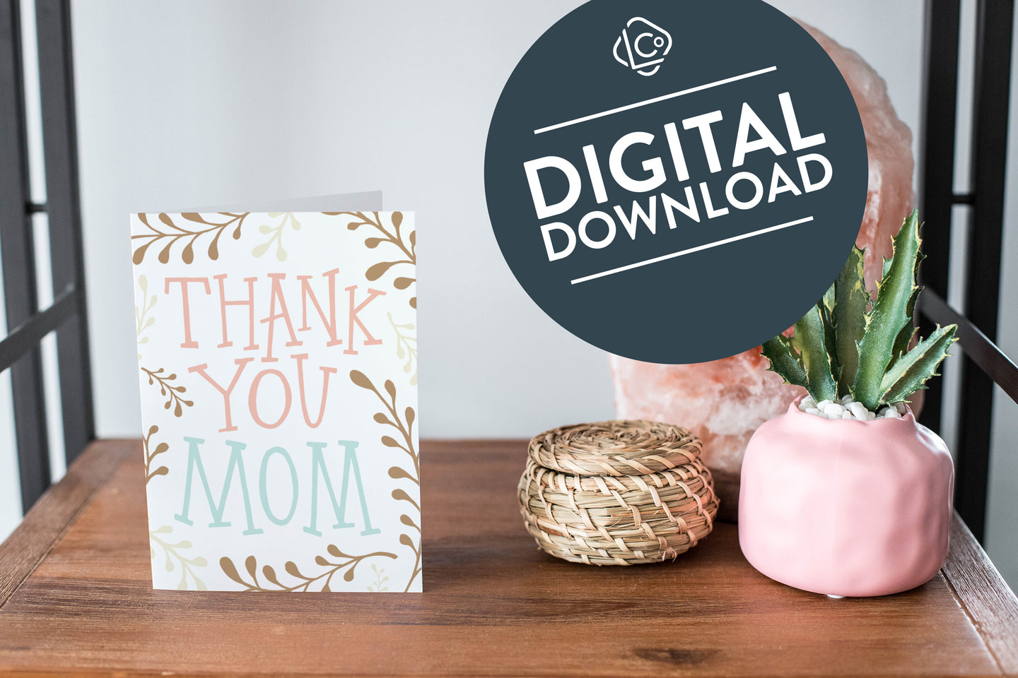 A card on a wood tabletop and on the right side of the card is a woven basket, a pink plant pot with a cactus in it and a pink crystal rock. The card features the words “Thank You Mom” with illustrated plant leaves. The words 