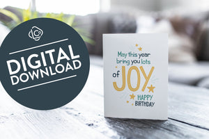 A greeting card is featured on a wood coffee table with a green plant in a white planter in the background. The card features the words “May This Year Bring You Lots of Joy Happy Birthday.”  The words "digital download" are featured in a circle on top of the image. 
