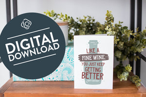 A greeting card is on a table top with a present in blue wrapping paper in the background. On top of the present is a candle and some greenery from a plant too. The card features the words "Like a fine wine, you keep getting better, Happy Birthday!” with an illustration of a wine bottle behind the words. The words "digital download" are featured in a circle on top of the image. 