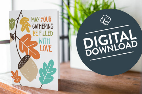A greeting card is featured on a wood coffee table with a green plant in a white planter in the background. The card features the words “May Your Gathering Be Filled with Love” with illustrated leaves and an acorn around the words. The words 