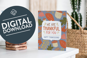 A greeting card featured standing up on a white tabletop with a stack of coasters next to it. There’s a woven basket in the background with a cactus inside. The card features the words "We are Thankful for You, Happy Thanksgiving" with illustrated leaves and acorns around the words. The words "digital download" are featured in a circle over the image.