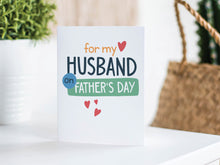 Load image into Gallery viewer, A greeting card is featured on a white tabletop with a white planter in the background with a green plant. There’s a woven basket in the background with a cactus inside. The card features the words “For my Husband on Father&#39;s Day” with small hearts around it. 