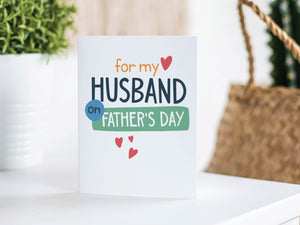 A greeting card is featured on a white tabletop with a white planter in the background with a green plant. There’s a woven basket in the background with a cactus inside. The card features the words “For my Husband on Father's Day” with small hearts around it. 