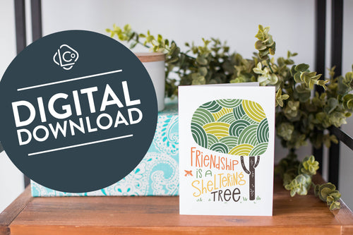 A greeting card is on a table top with a present in blue wrapping paper in the background. On top of the present is a candle and some greenery from a plant too. The card features the words “Friendship is a sheltering tree” with an illustrated tree. The words 