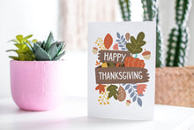 Load image into Gallery viewer, A greeting card featured standing up on a white tabletop with a pink plant pot with succulents. There’s a woven basket in the background with a cactus inside. The card features the words “Happy Thanksgiving” with illustrated leaves, a pumpkin, and acorn.