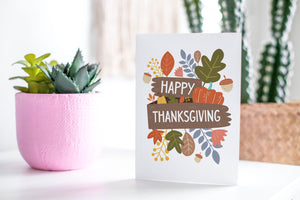 A greeting card featured standing up on a white tabletop with a pink plant pot with succulents. There’s a woven basket in the background with a cactus inside. The card features the words “Happy Thanksgiving” with illustrated leaves, a pumpkin, and acorn.