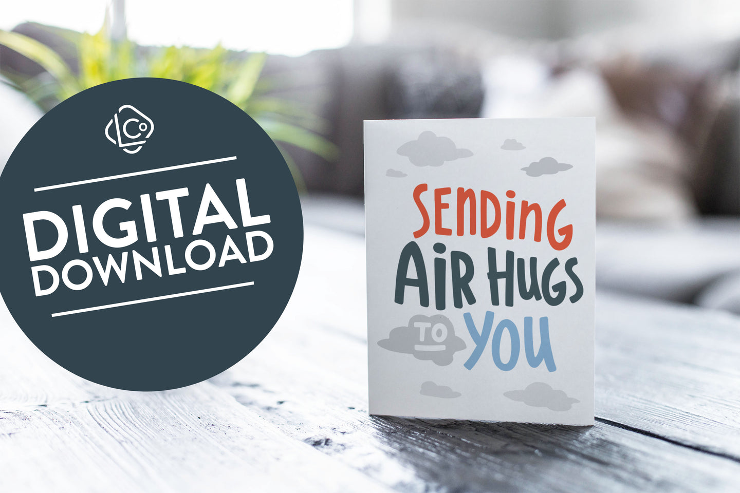 A greeting card featured on a black, wood coffee table. There’s a white planter in the background with a green plant. There’s also a gray sofa in the background with a white pillow. The card features the words “Sending air hugs to you.”. The words 