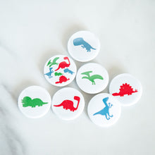 Load image into Gallery viewer, A set of seven buttons on a white tabletop. The buttons feature illustrations of a Pterodactyl, Sauropod, Stegosaurus, T-Rex, Triceratops, Velociraptor, and a button with all on one button. 