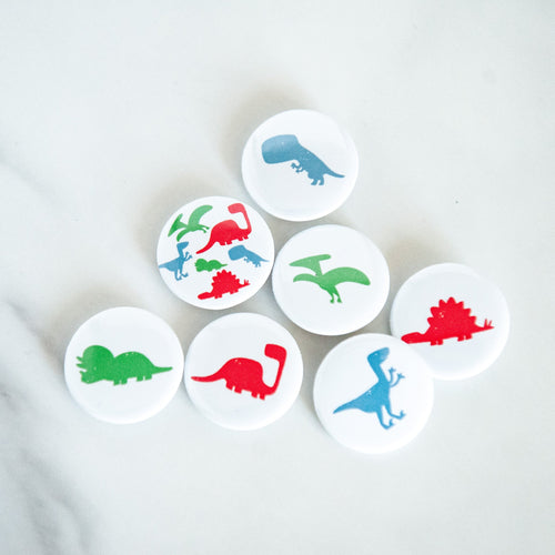 A set of seven buttons on a white tabletop. The buttons feature illustrations of a Pterodactyl, Sauropod, Stegosaurus, T-Rex, Triceratops, Velociraptor, and a button with all on one button. 