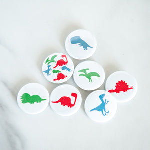 A set of seven buttons on a white tabletop. The buttons feature illustrations of a Pterodactyl, Sauropod, Stegosaurus, T-Rex, Triceratops, Velociraptor, and a button with all on one button. 