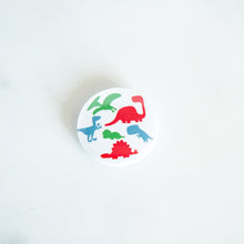 Load image into Gallery viewer, A button pin on a white tabletop. The button features an illustration of dinosaurs - Pterodactyl, Sauropod, Stegosaurus, T-Rex, Triceratops, and a Velociraptor. 