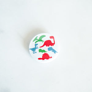 A button pin on a white tabletop. The button features an illustration of dinosaurs - Pterodactyl, Sauropod, Stegosaurus, T-Rex, Triceratops, and a Velociraptor. 