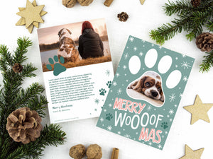 A photo of a two-sided Christmas card showing the front of the card on top of a brown wrapped gift on a white tabletop. Around the gift are pine needles, pinecones and wood star ornaments. The front of the card features a photo inside a paw shaped frame. Around the photo are white stars and below are the words “Merry Woofmas.” The back of the card features one photo with a dog paw illustration and space to add an update.