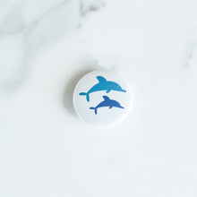 Load image into Gallery viewer, A button on a white tabletop. The button features an illustration of dolphins. 