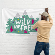 Load image into Gallery viewer, A man hanging a white flag on a wall. The white flag features the words &#39;wild and free&#39; inside illustrated mountains with sketch trees and other elements around.