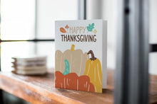 Load image into Gallery viewer, A card on a wood tabletop with an object in the background that is out of focus. The card features the words &quot;Happy Thanksgiving” with illustrated pumpkins below the words. 