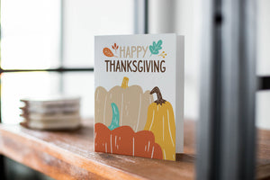 A card on a wood tabletop with an object in the background that is out of focus. The card features the words "Happy Thanksgiving” with illustrated pumpkins below the words. 