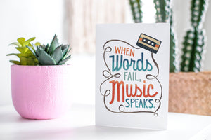 A greeting card featured standing up on a white tabletop with a pink plant pot in the background and some succulents in the pot. There’s a woven basket in the background with a cactus inside. The card features the words “When words fail, music speaks.”