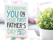 Load image into Gallery viewer, A greeting card is on a table top with a gift in pink wrapping paper. Next to the gift is a white plant pot with a green plant. The card features the words &quot;Celebrating you on your first Father&#39;s Day&quot; in modern, simple lettering with a coffee mug with #1 Dad on the mug.