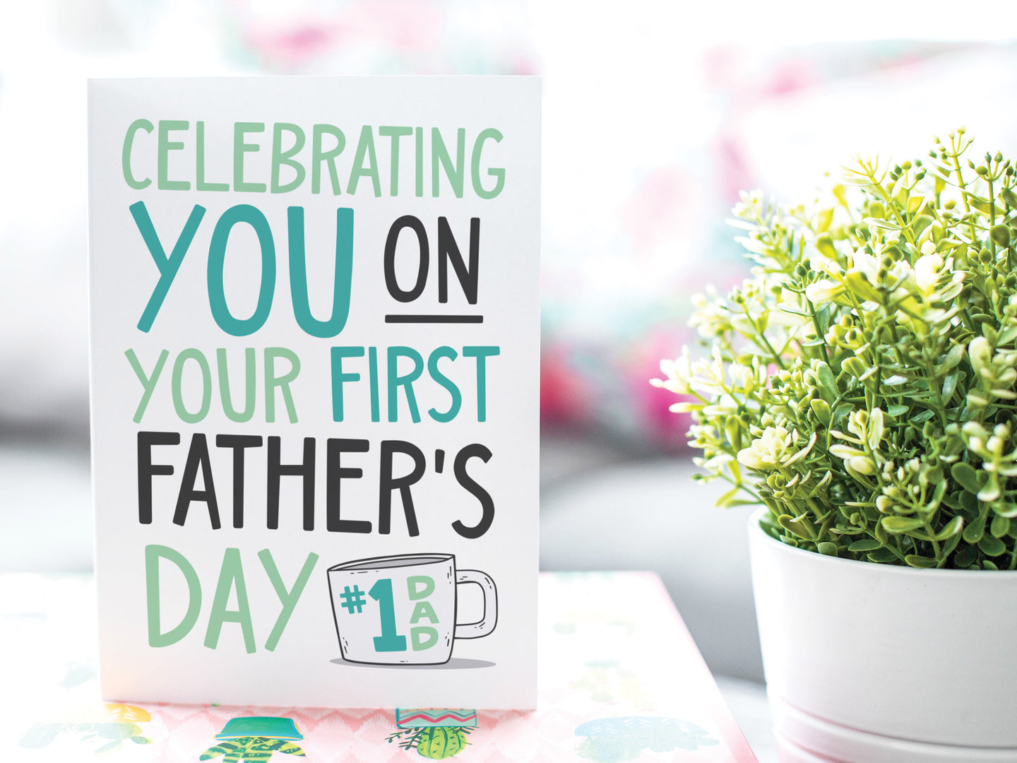 A greeting card is on a table top with a gift in pink wrapping paper. Next to the gift is a white plant pot with a green plant. The card features the words 