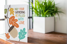 Load image into Gallery viewer, A greeting card is featured on a wood coffee table with a green plant in a white planter in the background. The card features the words “May Your Gathering Be Filled with Love” with illustrated leaves and an acorn around the words.