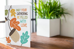 A greeting card is featured on a wood coffee table with a green plant in a white planter in the background. The card features the words “May Your Gathering Be Filled with Love” with illustrated leaves and an acorn around the words.