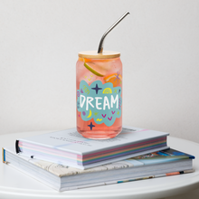 Load image into Gallery viewer, A photo of a glass can tumbler on a coffee table on top of books. The glass tumbler has a bamboo lid and silver metal straw. The design on the glass tumbler says the word &quot;dream&quot; inside a teal cloud illustration with sketchy illustrations around. 