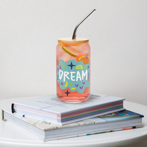 A photo of a glass can tumbler on a coffee table on top of books. The glass tumbler has a bamboo lid and silver metal straw. The design on the glass tumbler says the word "dream" inside a teal cloud illustration with sketchy illustrations around. 