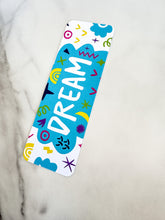 Load image into Gallery viewer, A bookmark on a tabletop. The bookmark design has the word &quot;dream&quot; inside a teal cloud-like shape with sketchy, whimsical items around. 