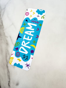 A bookmark on a tabletop. The bookmark design has the word "dream" inside a teal cloud-like shape with sketchy, whimsical items around. 