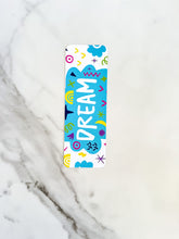 Load image into Gallery viewer, A bookmark on a tabletop. The bookmark design has the word &quot;dream&quot; inside a teal cloud-like shape with sketchy, whimsical items around. 