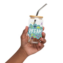 Load image into Gallery viewer, A photo of a glass can tumbler being held in a hand. The glass tumbler has a bamboo lid and silver metal straw. The design on the glass tumbler says the word &quot;dream&quot; inside a teal cloud illustration with sketchy illustrations around. 