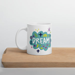 A white mug on a wood board. The mug features a design with the word 'dream' in an illustrated cloud with sketchy elements around it. 