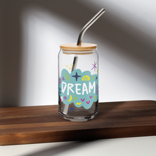 Load image into Gallery viewer, A photo of a glass can tumbler on a dark wood tabletop. The glass tumbler has a bamboo lid and silver metal straw. The design on the glass tumbler says the word &quot;dream&quot; inside a teal cloud illustration with sketchy illustrations around. 