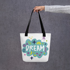 A person holding a white tote bag with black handles. The bag features a design with the word 'dream' with an illustrated cloud around the word with colorful sketchy around. 