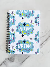 Load image into Gallery viewer, A spiral notebook is on a tabletop. The white spiral notebook has black spiral binding. The pattern design features the word &quot;dream&quot; inside a cloud shape with sketchy elements around. 