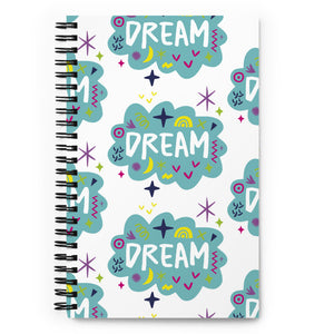 A spiral notebook is on a tabletop. The white spiral notebook has black spiral binding. The pattern design features the word "dream" inside a cloud shape with sketchy elements around. 