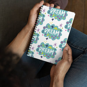 A spiral notebook being held by a person. The white spiral notebook has black spiral binding. The pattern design features the word "dream" inside a cloud shape with sketchy elements around. 