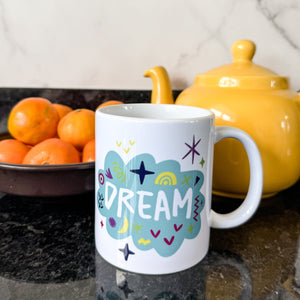 A white mug on a black countertop with a tea pot and oranges behind it. The mug features a design with the word 'dream' in an illustrated cloud with sketchy elements around it. 