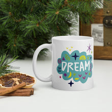 Load image into Gallery viewer, A white mug on a tabletop with part of a pine tree in the background. The mug features a design with the word &#39;dream&#39; in an illustrated cloud with sketchy elements around it. 