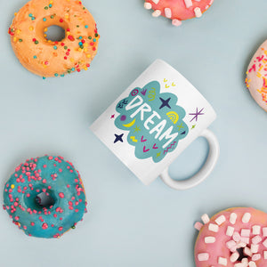 A white mug laying on a tabletop with doughnuts around it. The mug features a design with the word 'dream' in an illustrated cloud with sketchy elements around it. 